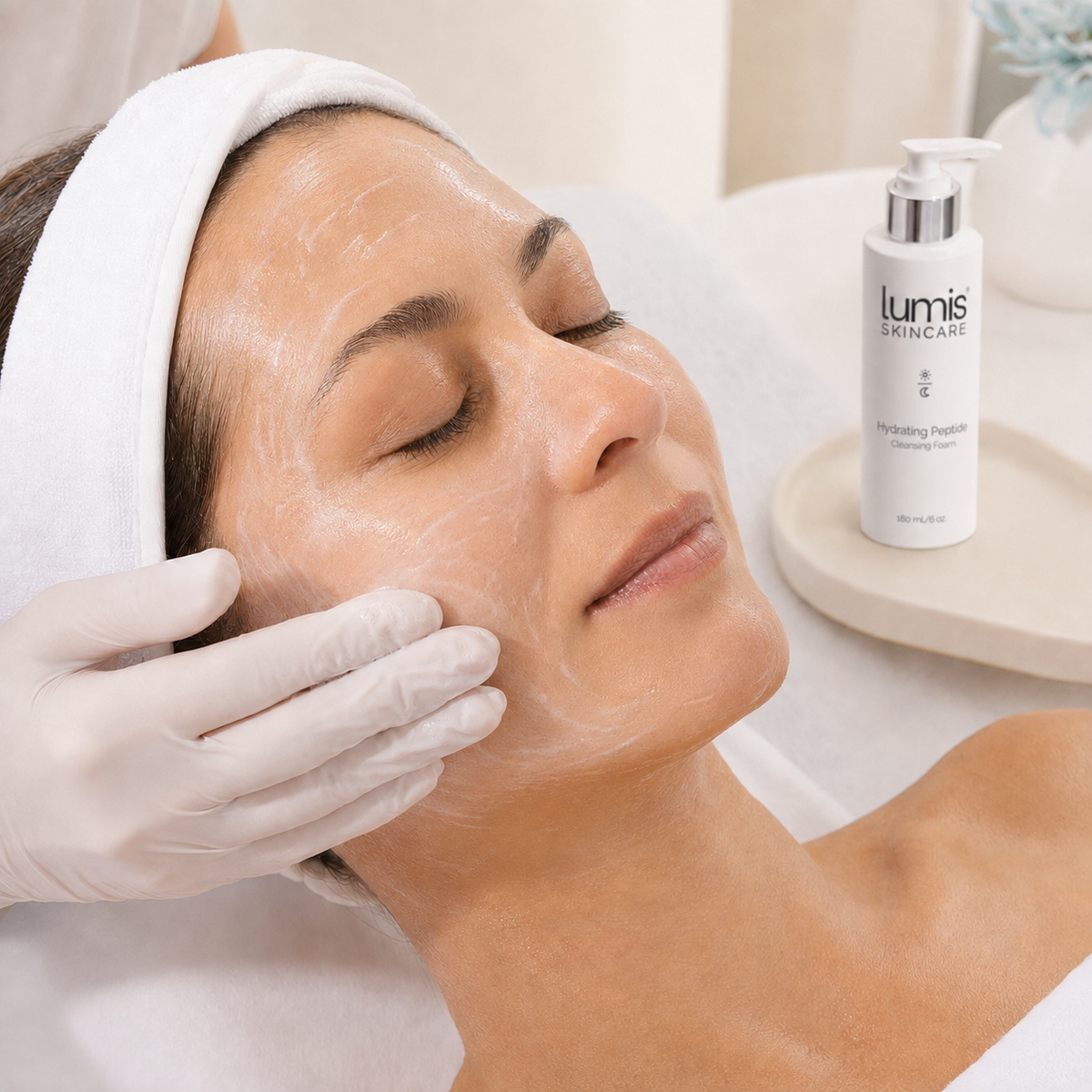 A woman receives a skincare treatment with a white cream on her face while wearing a white headband and gloves. A white pump bottle of Lumis SkinCare Hydrating Face Cream is visible on a small tray in the background.